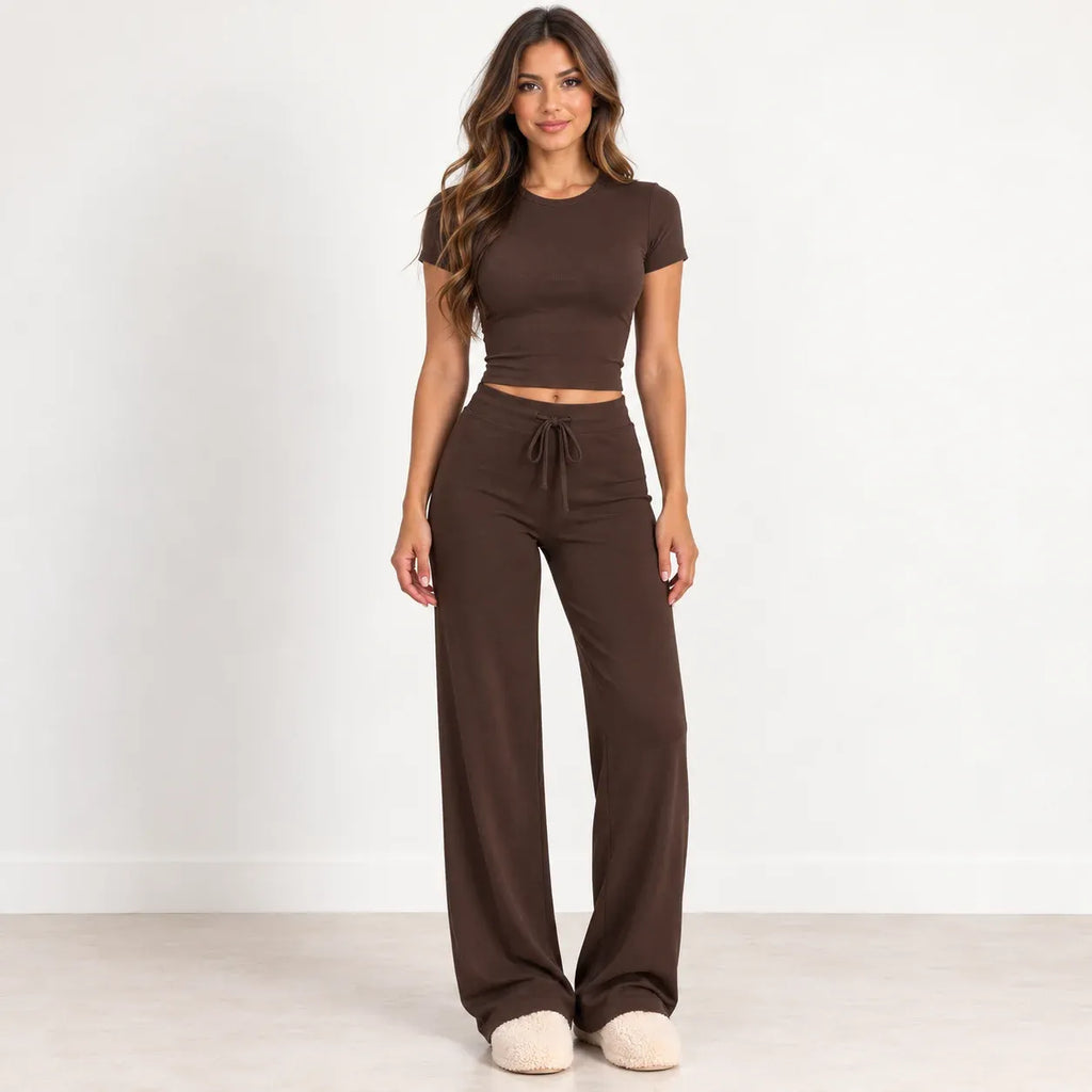 A woman modeling a matching two-piece lounge set consisting of a short-sleeve cropped t-shirt and high-waisted wide-leg drawstring pants in a solid dark brown color, paired with cream-colored slippers, set against a plain white studio background.