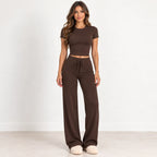 A woman modeling a matching two-piece lounge set consisting of a short-sleeve cropped t-shirt and high-waisted wide-leg drawstring pants in a solid dark brown color, paired with cream-colored slippers, set against a plain white studio background.