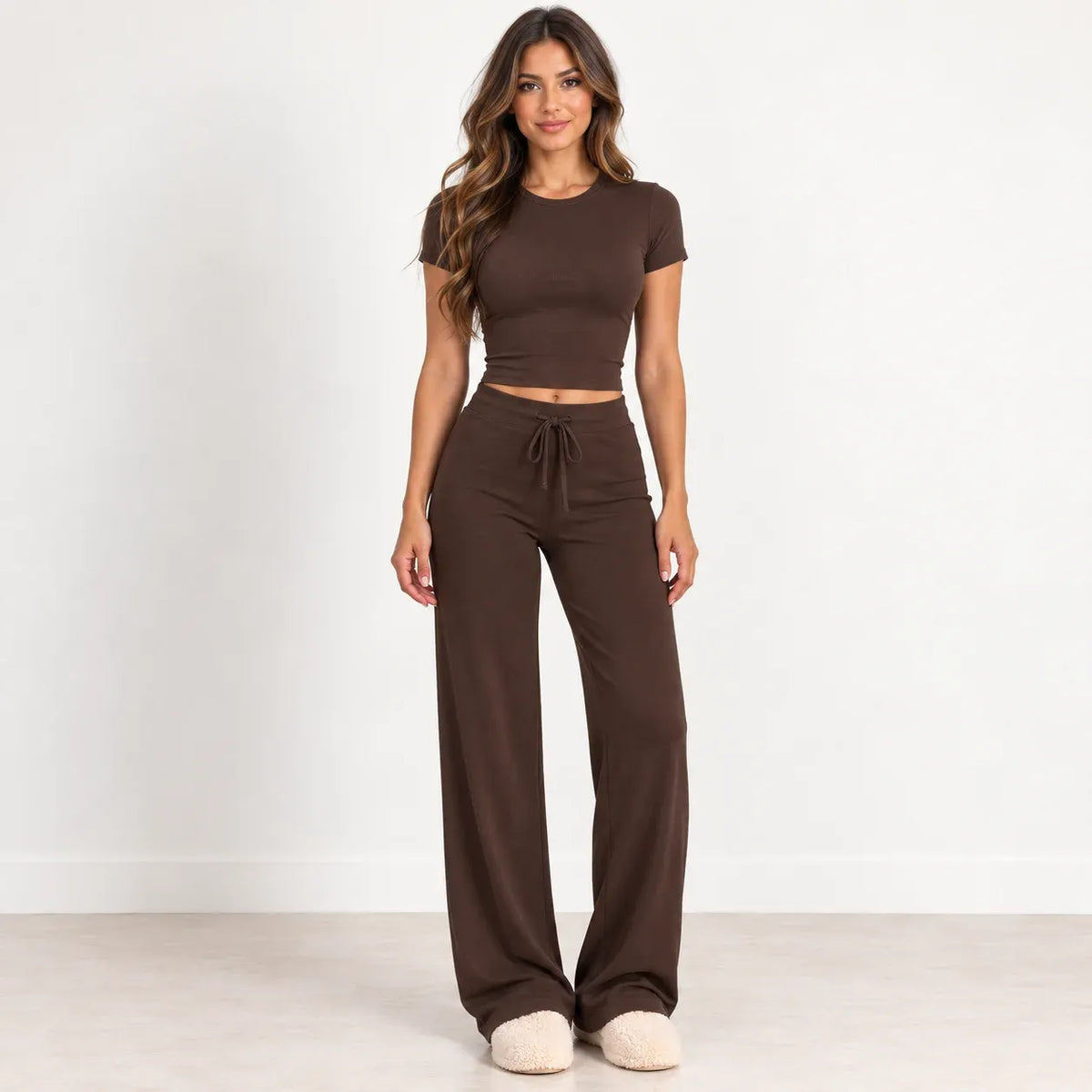 A woman modeling a matching two-piece lounge set consisting of a short-sleeve cropped t-shirt and high-waisted wide-leg drawstring pants in a solid dark brown color, paired with cream-colored slippers, set against a plain white studio background.