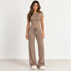 A woman modeling a matching two-piece lounge set in a taupe-brown color, featuring a short-sleeve crop top and high-waisted wide-leg pants with a drawstring waist, standing against a plain white studio background.
