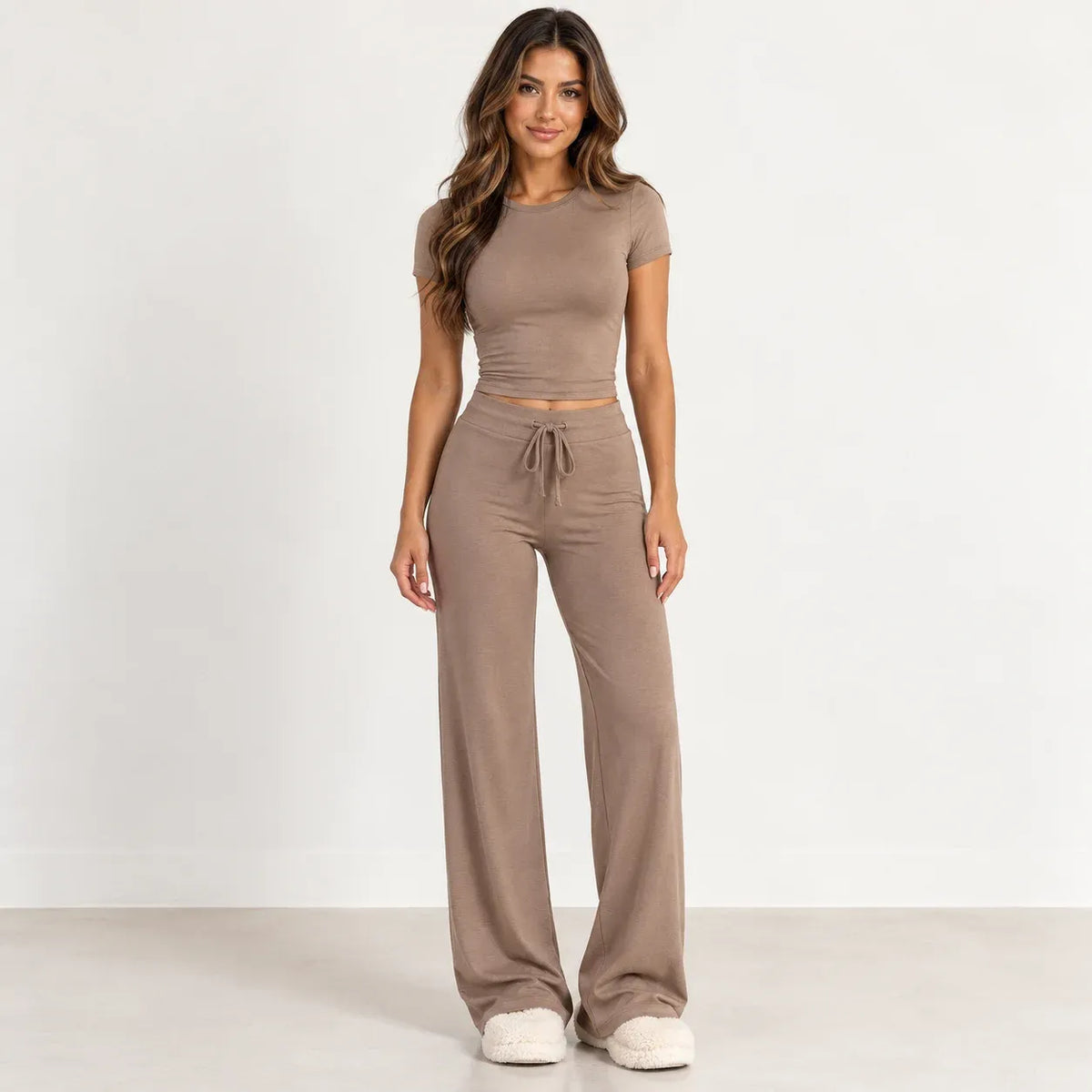 A woman modeling a matching two-piece lounge set in a taupe-brown color, featuring a short-sleeve crop top and high-waisted wide-leg pants with a drawstring waist, standing against a plain white studio background.
