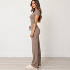 A woman stands in profile wearing a matching taupe two-piece loungewear set consisting of a short-sleeve crop top and high-waisted wide-leg pants, paired with cream fuzzy slippers against a neutral studio background.