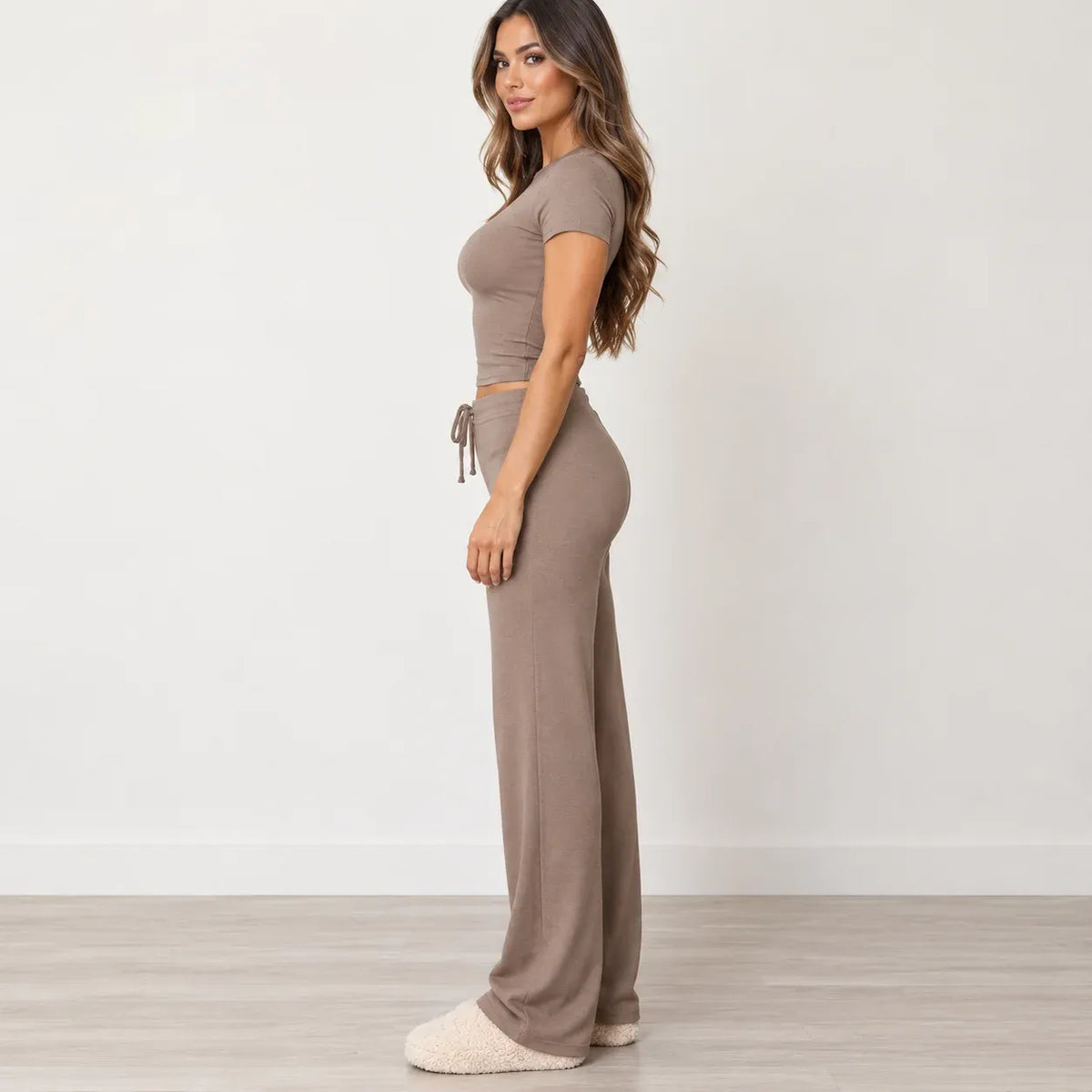A woman stands in profile wearing a matching taupe two-piece loungewear set consisting of a short-sleeve crop top and high-waisted wide-leg pants, paired with cream fuzzy slippers against a neutral studio background.