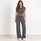 A woman modeling a dark gray two-piece loungewear set consisting of a short-sleeve crop top and matching wide-leg drawstring pants, standing against a white wall.