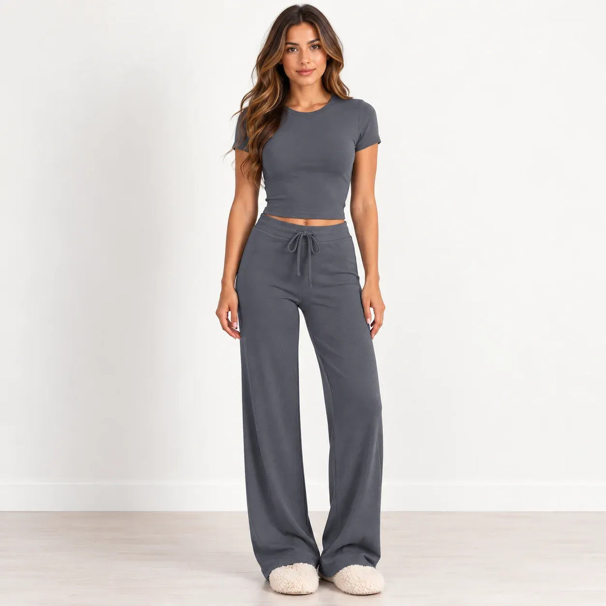 A woman modeling a dark gray two-piece loungewear set consisting of a short-sleeve crop top and matching wide-leg drawstring pants, standing against a white wall.
