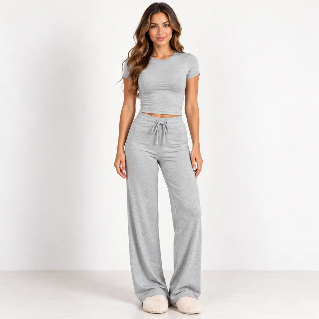 A woman modeling a light gray two-piece loungewear set consisting of a short-sleeve crop top and matching wide-leg drawstring pants, standing against a plain white studio background.
