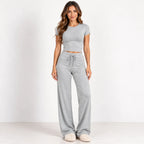A woman modeling a light gray two-piece loungewear set consisting of a short-sleeve crop top and matching wide-leg drawstring pants, standing against a plain white studio background.