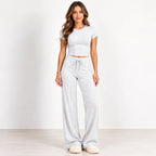 A woman modeling a light gray two-piece loungewear set consisting of a short-sleeve crop top and matching wide-leg sweatpants, standing against a plain white studio background.