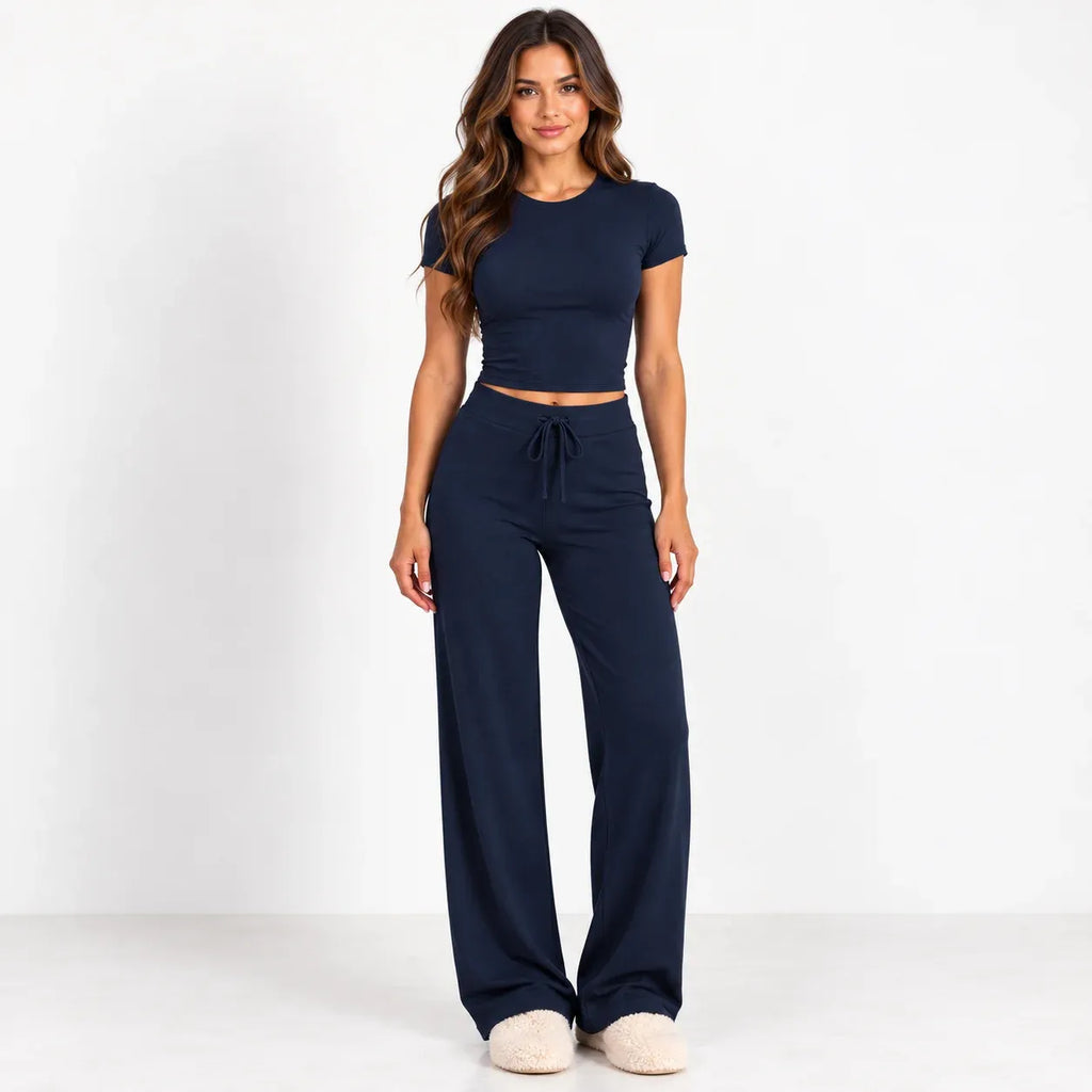 A woman modeling a matching navy blue two-piece loungewear set, consisting of a short-sleeve crop top and high-waisted wide-leg pants with a drawstring waist, standing against a plain white studio background.