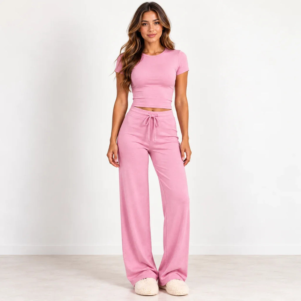 A woman modeling a pink two-piece loungewear set consisting of a short-sleeve crop top and matching wide-leg drawstring pants, standing against a plain white studio wall.