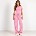 A woman modeling a pink two-piece loungewear set consisting of a short-sleeve crop top and matching wide-leg drawstring pants, standing against a plain white studio wall.