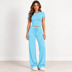 A woman modeling a light blue two-piece loungewear set consisting of a short-sleeve crop top and matching wide-leg drawstring pants, standing against a plain white wall.