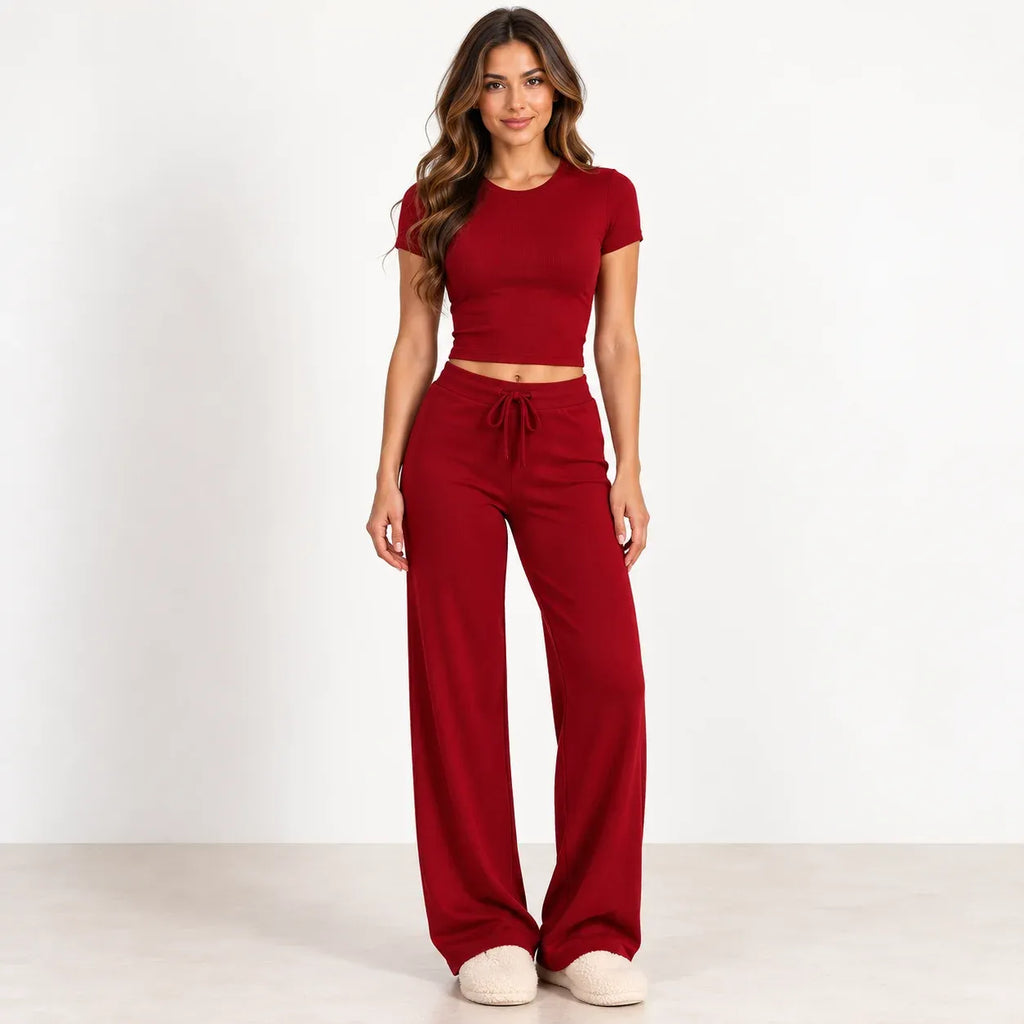 A woman modeling a matching two-piece loungewear set consisting of a short-sleeve crop top and high-waisted wide-leg pants in a deep red color, paired with beige slippers against a plain white studio background.