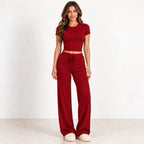 A woman modeling a matching two-piece loungewear set consisting of a short-sleeve crop top and high-waisted wide-leg pants in a deep red color, paired with beige slippers against a plain white studio background.