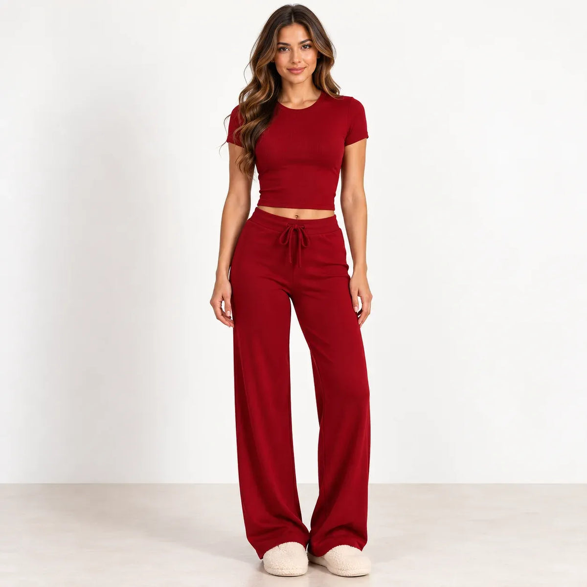 A woman modeling a matching two-piece loungewear set consisting of a short-sleeve crop top and high-waisted wide-leg pants in a deep red color, paired with beige slippers against a plain white studio background.