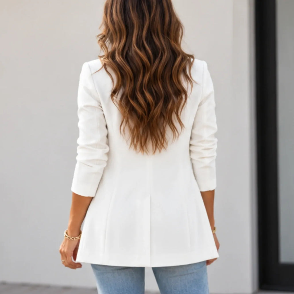 A woman viewed from behind wearing a white long-sleeve blazer with a peplum-like flare at the hem, paired with light blue jeans. She has long, wavy brown hair and is wearing a gold bracelet.