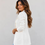 A woman with long wavy brown hair is shown from the side and back, wearing a white blazer over a light blue top. She is standing against a plain light gray wall.