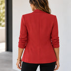 A woman viewed from the back wearing a structured red blazer with three-quarter sleeves and black pants, standing indoors against a light neutral wall.