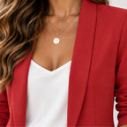 A close-up of a woman wearing a red blazer over a white top, accessorized with layered gold necklaces, including a round disc pendant.