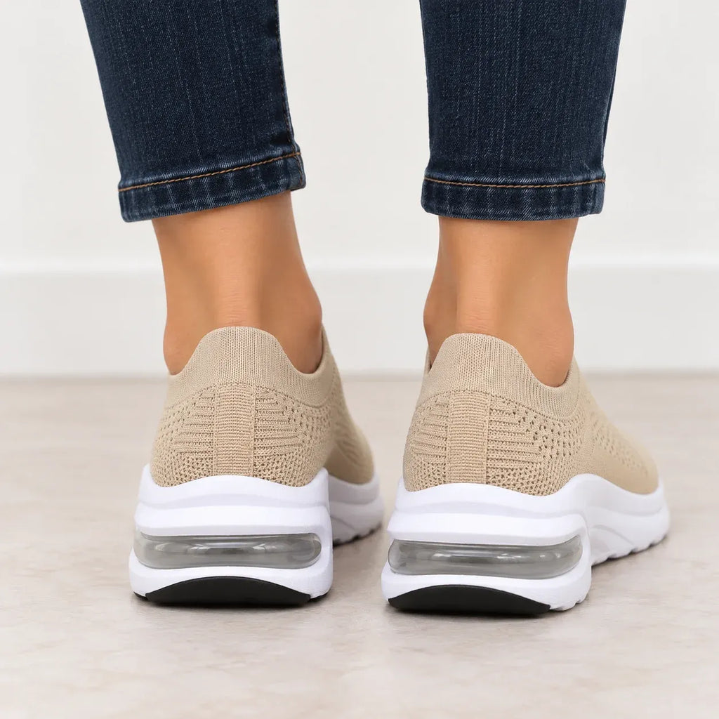 A back view of a pair of beige slip-on knit sneakers with white soles and clear air cushioning, shown on a person wearing blue jeans.