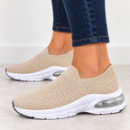 A pair of beige slip-on knit sneakers with white soles and clear air cushioning, shown on a person wearing blue jeans.
