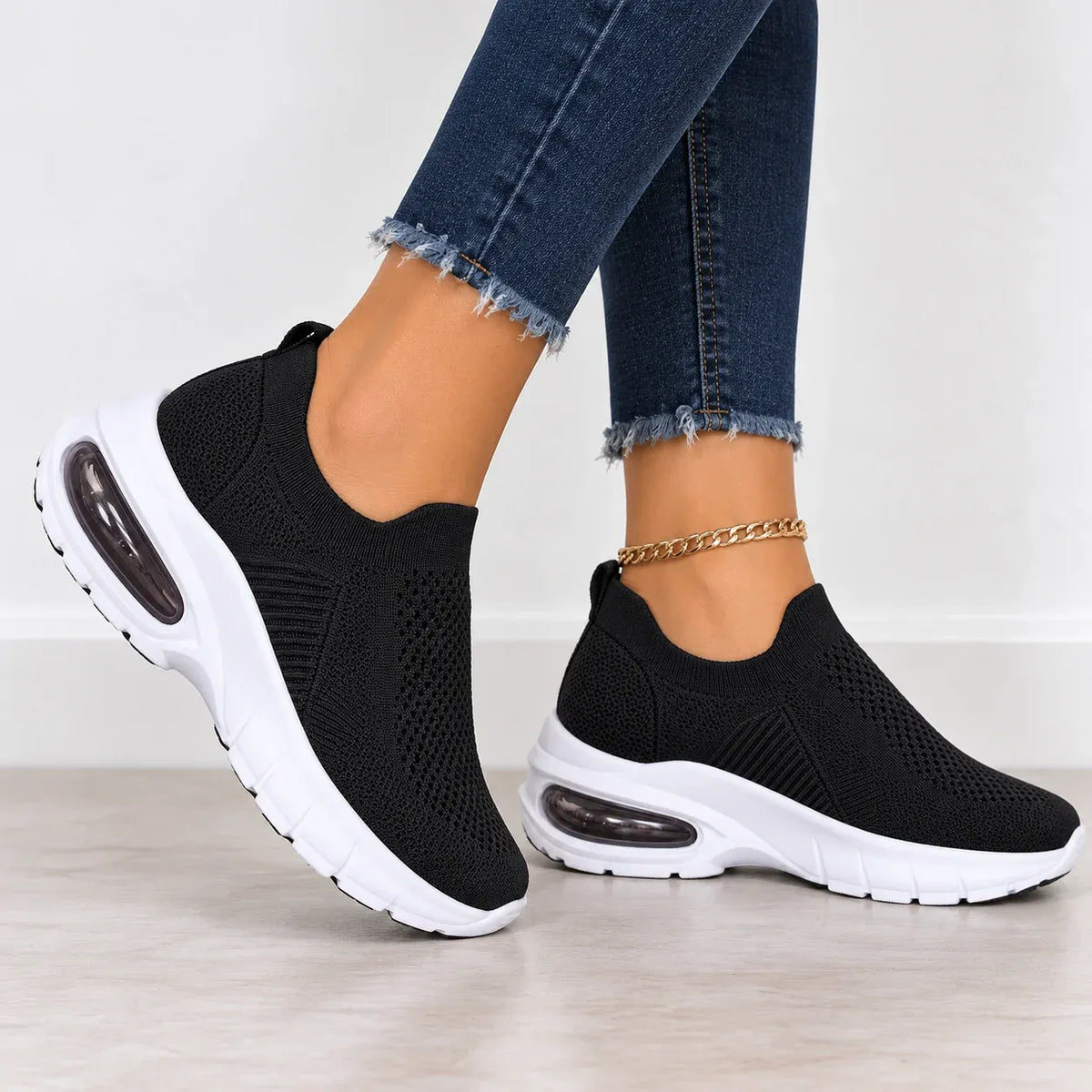 A pair of black slip-on knit sneakers with chunky white soles and visible air cushioning, shown on a model's feet wearing frayed blue jeans against a light indoor background.