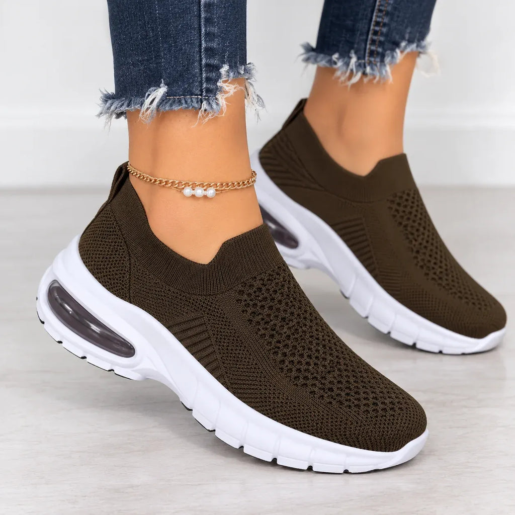 A pair of women's slip-on knit sneakers in dark brown with white soles, shown on a model's feet wearing frayed blue jeans.