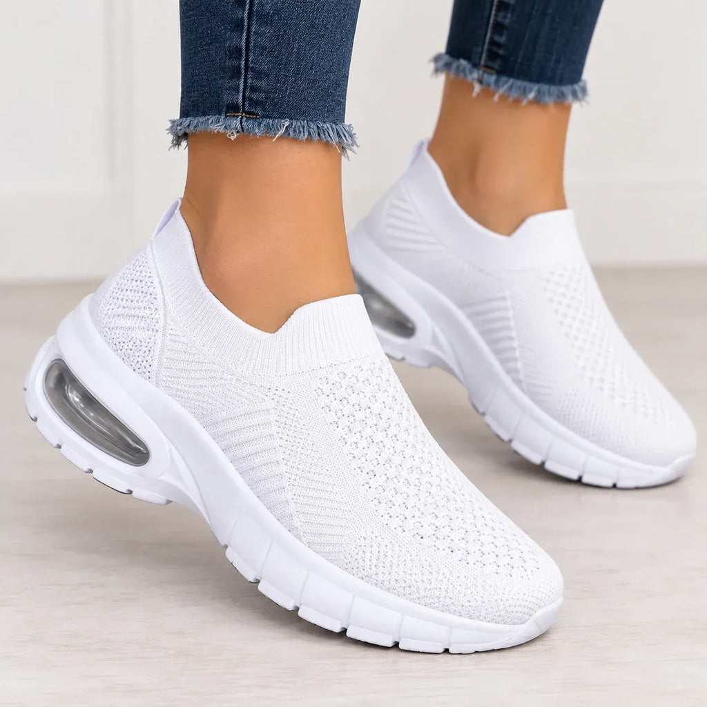 White slip-on knit sneakers with a visible air cushion unit in the sole, shown on a person's feet wearing frayed blue jeans.
