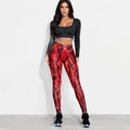 A woman modeling high-waisted red and black snakeskin-patterned leggings with a black long-sleeve crop top and black sneakers, standing against a plain white studio wall.