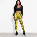 A woman modeling high-waisted leggings with a bold yellow and black snakeskin pattern, paired with a black long-sleeve crop top and black sneakers, standing against a plain white wall.