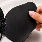 A close-up of a black lace-up bra cup being held by a hand with light pink manicured nails, showing the matte texture of the cup and part of the black lace-up front.