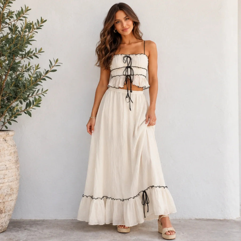 A woman models a two-piece cream maxi set featuring a ruffled crop top with black lace-up details and a matching long skirt with a ruffled hem and black tie accents, set against a white wall with a potted olive tree on the left.