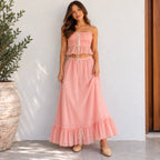 A woman modeling a two-piece pink bohemian-style outfit consisting of a ruffled crop top with tie details and a matching long ruffled maxi skirt, standing against a white wall next to a potted plant.