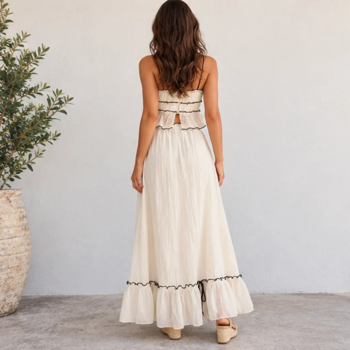 A woman viewed from behind wearing a strapless cream maxi dress with black lace trim and a ruffled back detail, standing against a light gray wall next to a potted plant.