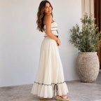 A woman wearing a long white maxi dress with black trim and side ties, standing in a bright indoor setting with a large potted plant in the background.