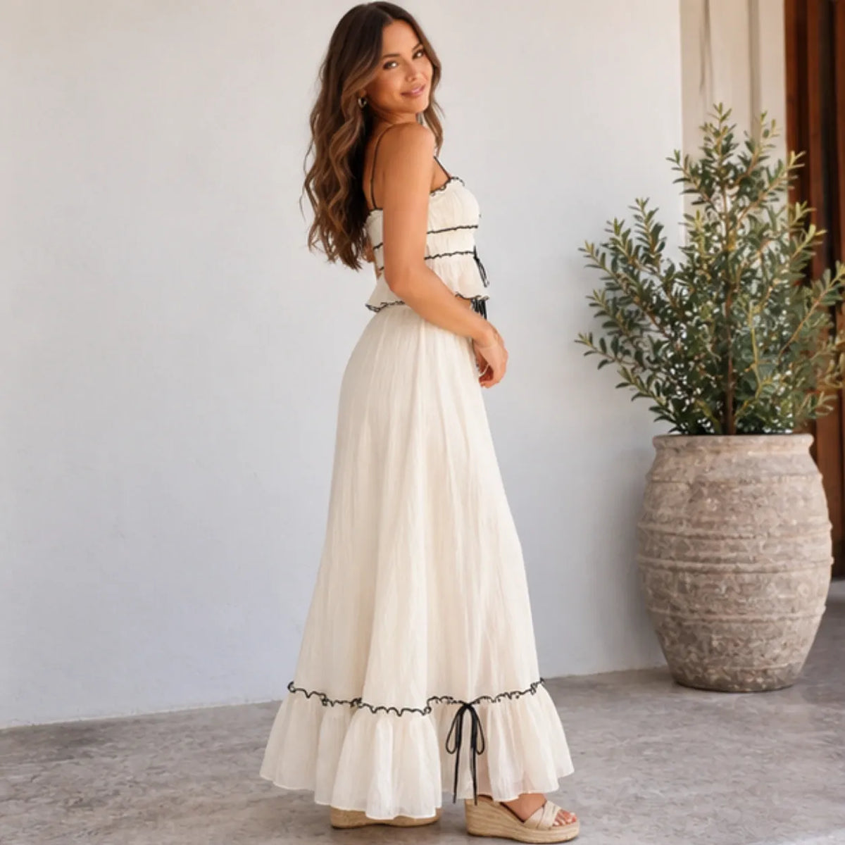 A woman wearing a long white maxi dress with black trim and side ties, standing in a bright indoor setting with a large potted plant in the background.