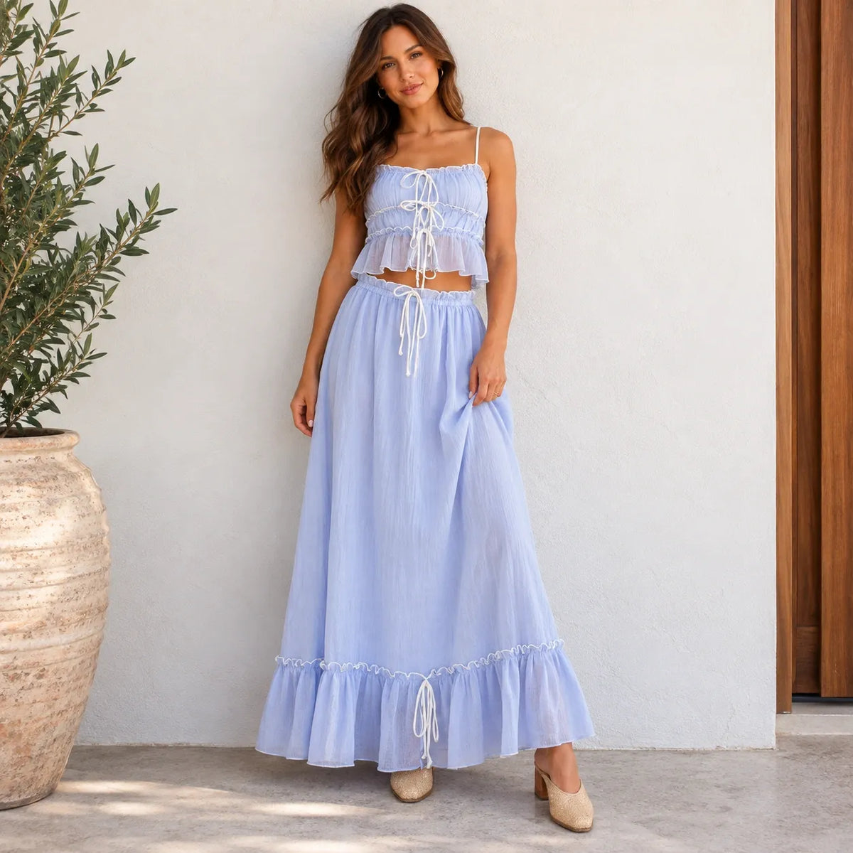 A woman models a light blue two-piece maxi dress set consisting of a crop top with white tie details and a matching high-waisted maxi skirt with a ruffled hem, standing against a white stucco wall next to a potted olive tree.