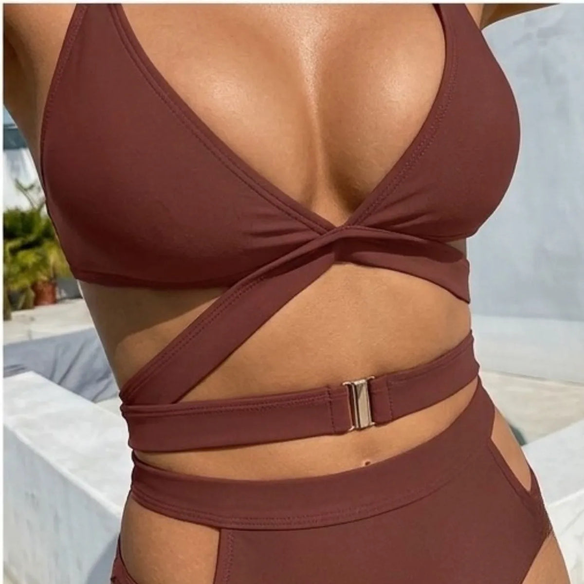 A close-up of a model wearing a deep burgundy two-piece swimsuit with a crossover halter bikini top and matching high-waisted bottoms featuring a side cutout and a silver buckle detail.