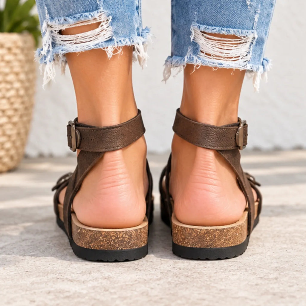 A close-up rear view of a person wearing dark brown strappy platform sandals with cork-look midsoles and black soles, paired with frayed light-wash denim jeans.
