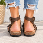 A close-up rear view of a person wearing dark brown strappy platform sandals with cork-look midsoles and black soles, paired with frayed light-wash denim jeans.