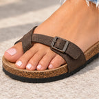 A close-up of a person's foot wearing a brown crisscross sandal with a buckle strap and a cork-textured sole, standing on a light concrete surface.
