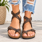 A pair of dark brown strappy wedge sandals with buckle details, shown on a model's feet wearing ripped blue jeans.