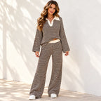 A woman modeling a matching two-piece knit set with horizontal black and cream stripes, featuring a long-sleeve cropped polo-style top with a V-neck and wide-leg pants, standing against a white wall with soft shadows.