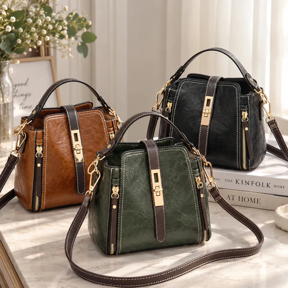 Three faux-leather handbags with structured rectangular shapes, top handles, and shoulder straps are arranged on a light marble surface. The bags are shown in brown, dark green, and black, each with gold-tone hardware and zipper details.
