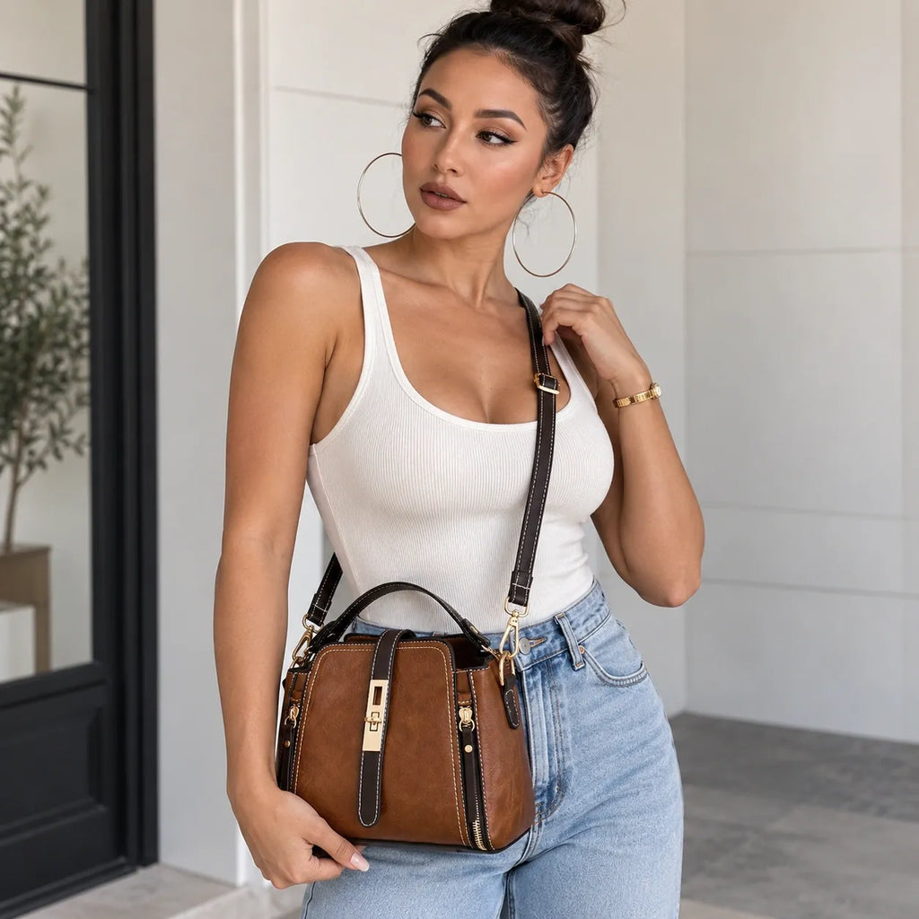 A woman wearing a white ribbed tank top and light blue jeans carries a brown leather crossbody bag with gold-tone hardware and a black strap, standing in a bright modern interior.