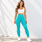 A woman modeling a matching activewear set consisting of a white sports bra and high-waisted textured teal leggings, standing against a white stucco wall.