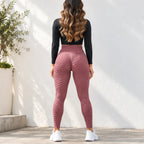 A woman is shown from behind wearing textured, high-waisted dusty rose leggings, a black long-sleeve crop top, and white sneakers, standing against a white stucco wall.