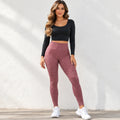 A woman modeling a two-piece activewear set consisting of a black long-sleeve crop top and textured dusty rose leggings, paired with white sneakers, standing against a white textured wall.