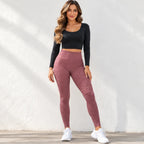 A woman modeling a two-piece activewear set consisting of a black long-sleeve crop top and textured dusty rose leggings, paired with white sneakers, standing against a white textured wall.