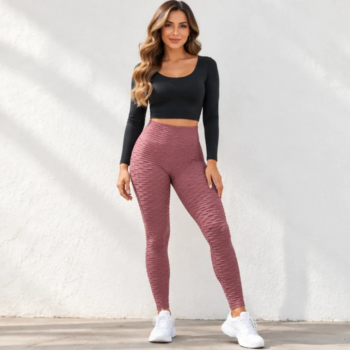 A woman modeling a two-piece activewear set consisting of a black long-sleeve crop top and textured dusty rose leggings, paired with white sneakers, standing against a white textured wall.